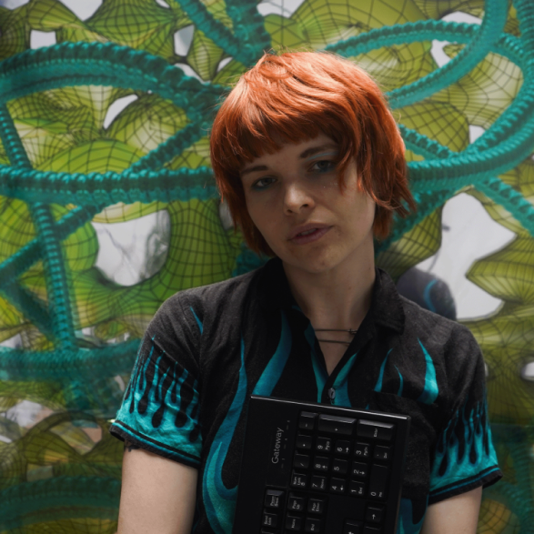 An image of Tee Topor, with red hair standing in front of a green pattern background, wearing a blue flame shirt and keyboard