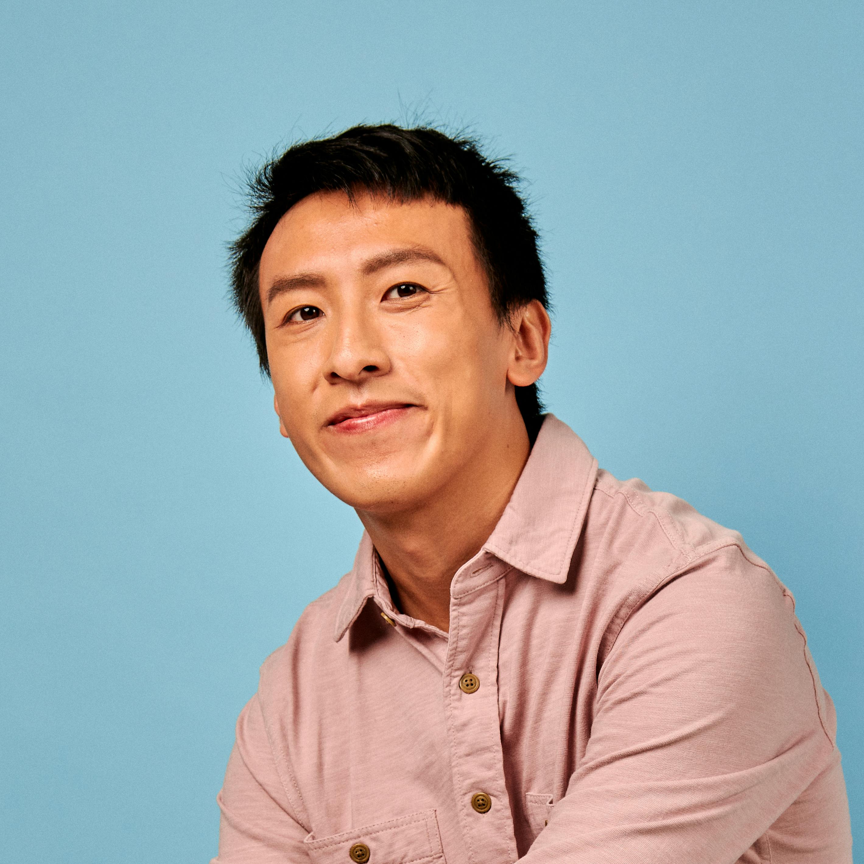 A man of southeast-asian descent smiles in front of blue background.